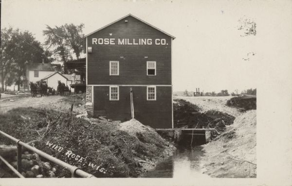 Text on front reads: "Wild Rose, Wis." View from bridge, with a railing in the foreground. There are two horse-drawn wagons standing next to a mill on the left near two men standing on the loading dock. Behind them is a dwelling. "Rose Milling Co." is painted on the side of the mill. The dam, on the Pine River, is located on the right. The mill was originally built in 1873, and redeveloped in 1904. A water-powered generator was added in 1908. The property was purchased by the village and the dam was reconstructed.