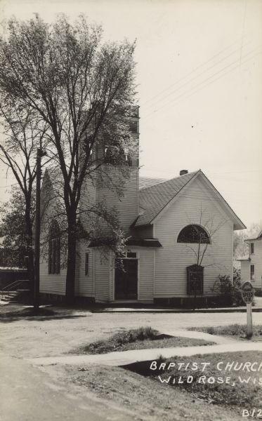 Text on front reads: "Baptist Church, Wild Rose, Wis." Built in 1901 of clapboard in the Queen Anne style. The street is unpaved, and there are sidewalks and trees. In 2007 the congregation moved to a new church in a new location. The bell and several stained glass windows were incorporated into the new building.