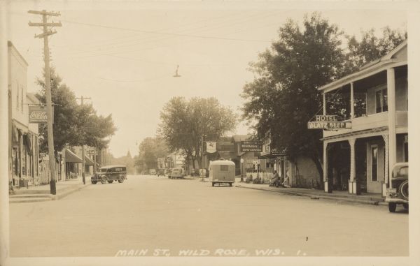 Text on front reads: "Main St., Wild Rose, Wis." Two women are sitting on a bench on the sidewalk under the awning of a restaurant, a trailer is parked at the curb, and automobiles and a "GrayCo Service" truck are parked diagonally. The street is lined with businesses and trees. Signs read: "Drug Store, Coca-Cola", "Bake-Rite Bread Sold Here", "At Market Groceries", "Standard Service", "Schlitz", "Mobilgas", "Restaurant Home Cooking", "Hotel"and "Blatz Beer."