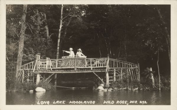 Text on front reads: "Long Lake. Woodlands. Wild Rose, Wis." A man and women are seated on a bridge-like platform over an inlet on Long Lake. The platform is built of logs and branches, with decorative designs in the railings. The background is filled with trees.<p>Long Lake is Northeast of Wild Rose, closer to Saxeville.</p>