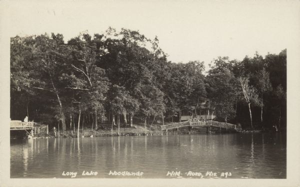 Text on front reads: "Long Lake. Woodlands. Wild Rose, Wis." View across lake towards a bridge-like platform in front of a cottage on Long Lake. The platform is built of logs and branches, with decorative designs in the railings. On the left is a similar structure with a person standing on it. The shoreline is filled with trees.<p>Long Lake is Northeast of Wild Rose, closer to Saxeville.</p>