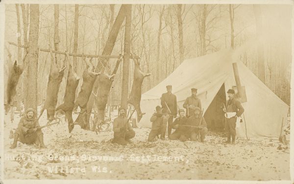Caption reads: "Hunting Scene, Slevenic [sic] Settlement, Willard, Wis." Group portrait of eight hunters with guns, and six deer carcasses hanging from a horizontal pole. On the right is a wall tent with a wood stove inside. Trees are in the background. Willard in Clark County had the only Slovenian agricultural community in Wisconsin.