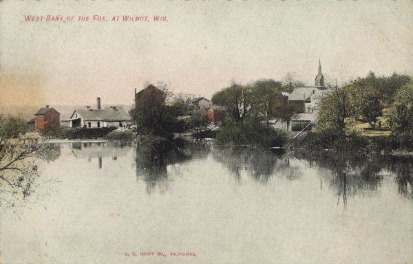 Text on front: "West Bank of the Fox, at Wilmot, Wis." View of a town from the Fox River. Trees and buildings are along the bank. A church steeple is in the distance. 