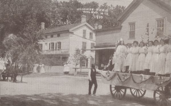 Text on the front reads: "Woodmen Picnic, Wilmot, Wis. 1910" On reverse: "Woodman Picnic – 1910, Wilmot, Wisconsin." A parade with horse-drawn wagons carrying women in light colored dresses. The woman on the end of the wagon on the right is wearing a crown, with her hair worn loose, and a more ornate gown. The wagon is decorated with bunting and flowers, and a man is standing on the ground at the rear. Another wagon is coming into view on the left. In the background spectators are standing on the sidewalk in front of two large buildings. Tree are in the background.