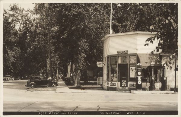 Text on front reads: "Post Office and Store, Winnebago, Wis." A corner store and post office with three gas pumps, surrounded by trees. A man leans on his parked automobile on the left. Three children can be seen on the sidewalk in the distance. Many signs read: "Carver Ice Cream", "Winnebago, Wis. Post Office", "Coca-Cola", "Dad's", "7-up", "Denver Sandwich 5¢", "Copenhagen", "Standard Red Crown", "Ringling Bros Barnum & Bailey", and "Oshkosh Aug 12." A "Wings Over America. Air Corps U.S. Army" poster hangs in the window.