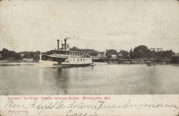 Text on front reads: "Steamer 'Le Fevre' Coming Through Bridge, Winneconne, Wis." A steamer passes through the "lost through truss swing bridge" over the Wolf River. Buildings and trees are on the shoreline.