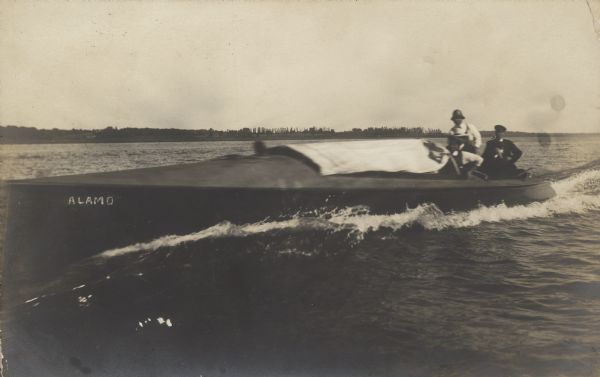 An early motor boat, named "Alamo," possibly on Lake Winneconne. Three men are in the passenger compartment. Buildings and trees are visible on the far shore.