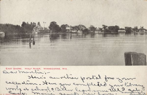 Text on front reads: "East Shore, Wolf River, Winneconne, Wis." Pilings in the Wolf River, boathouses, dwellings and trees are along the shore. A steamboat is on the far right.