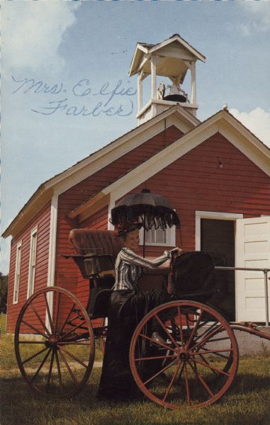 Text on the reverse reads: "The Fern Dell County School at The Pioneer Village Museum at Wisconsin Dells, Wis. Built in 1883 and used every year until 1955. Preserved in memory of the one room country schools and their teachers." Handwritten on the front: "Mrs. Elfie Farber." A fashionably dressed woman, holding a black parasol, is posing with her foot on the frame of a horse-drawn buggy. Behind is red clapboard one-room schoolhouse with white trim and a belfry.