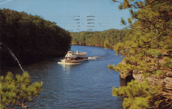 Text on reverse reads: "Cruising Down the River. A peaceful cruise plus the wonder found along its pine-covered, rocky shoreline make the boat trip at the Dells complete. Wisconsin Dells, Wis." A Wisconsin Dells tour boat on the Wisconsin River, surrounded by trees and rock formations.