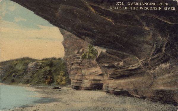 Text on front reads: "Overhanging Rock, Dells of the Wisconsin River." View of the Wisconsin River from under a rock shelf. The shoreline in the foreground is sandy. A small boat is on the shore in the distance.