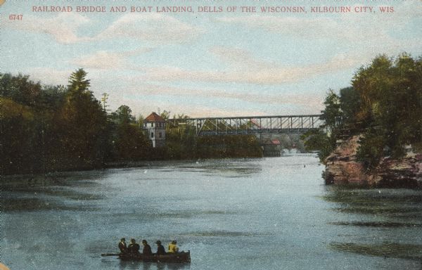 Text on front reads: "Railroad Bridge and Boat Landing, Dells of the Wisconsin, Kilbourn City, Wis." A bridge is in the background, which goes over the Wisconsin River. In the foreground are five people in a rowboat Buildings can be seen on the left near the bridge, and a rock formation is on the right. The bridge was built in 1877 as a Whipple deck truss span, replacing a wooden bridge. In 1903, a new bridge was built after it was destroyed by a tornado.<p>Kilbourn City was founded in 1857 by Byron Kilbourn, and the name was changed in 1931 to Wisconsin Dells.</p>