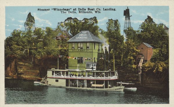 Text on front reads: "Steamer 'Winnebago' at Dells Boat Co. Landing, The Dells, Kilbourn, Wis." The steamboat "Winnebago," with passengers, at the Dells Boat Landing on the Wisconsin River. A building and trees are perched on the rock formation on the shoreline of the river with more buildings in the background and to the right.<p>Kilbourn City was founded in 1857 by Byron Kilbourn, the name was changed in 1931 to Wisconsin Dells.</p>