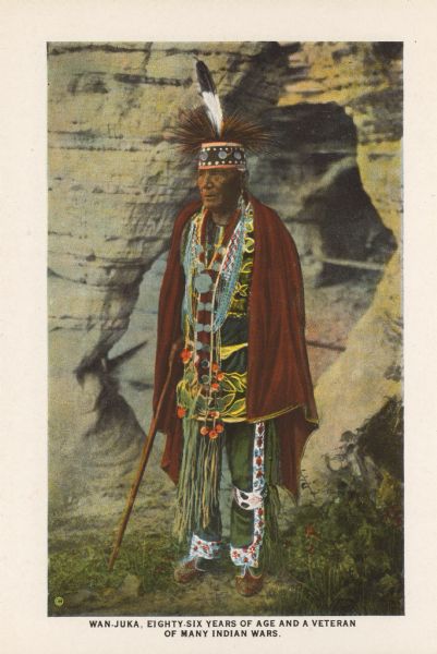 Text on front reads: "Wan-Juka, Eighty-Six Years of Age and a Veteran of Many Indian Wars." One of 16 postcard images inside of a souvenir view folder. A Native American man wearing indigenous clothing and carrying a cane. In the background is a rock formation. 