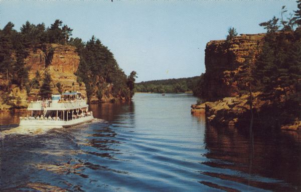 Text on reverse reads: "Wisconsin Dells, Wis. Clipper 'Winnebago' at the Lower Jaws of the Dells showing Alligator Rock on the upper right hand corner." A tour boat full of visitors on the Wisconsin River. Rock formations and trees line the river, a small boat can be seen in the distance.