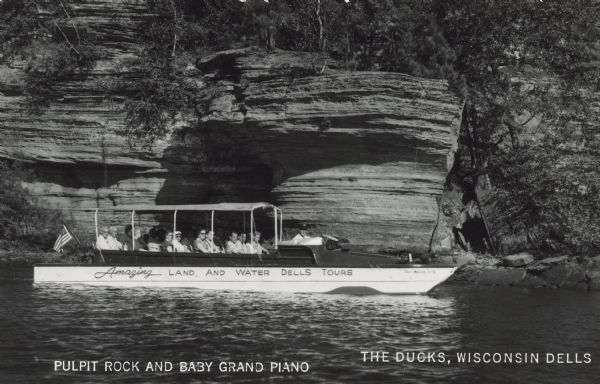 Text on front reads: "Pulpit Rock and Baby Grand Piano. The Ducks, Wisconsin Dells." Text on the "Duck" reads: "Amazing Land and Water Dells Tours." The name of the boat is "Chief Walking Turtle." A group of visitors in a Duck tour boat. Ducks are amphibious vehicles used during World WII for the transportation of goods and troops over land and water. The first Original Dells Duck Tour took place in 1946.