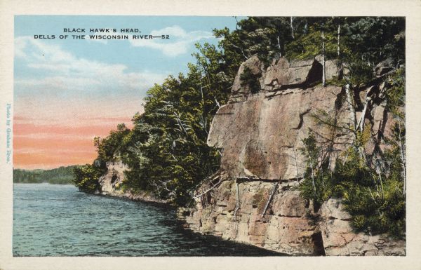 Text on front reads: "Black Hawk's Head, Dells of the Wisconsin River." On reverse: "Up stream, after passing High Rock and Chimney Rock, on the right hand bank is the wonderfully realistic Black Hawk's Profile, a greenish colored rock with a high slanting forehead and roman nose." A rock formation on the shore of the tree lined Wisconsin River.