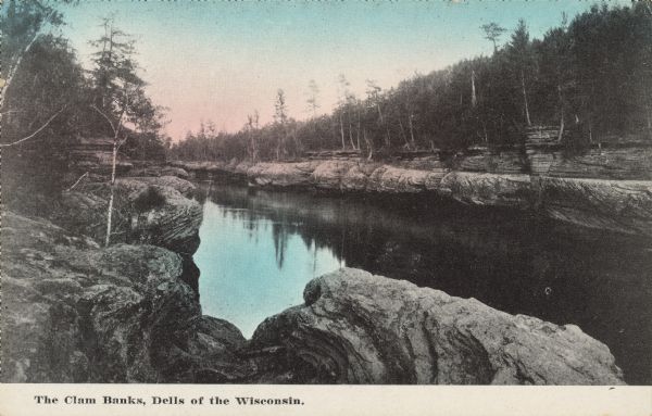 Text on front reads: "The Clam Banks, Dells of the Wisconsin." Rock formations called "The Clam Banks" for their resemblance to overlapping clam shells. The shore above the formations are filled with trees.
