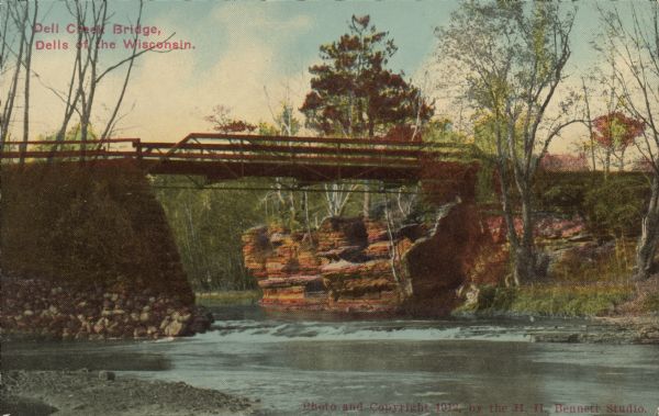 Text on front reads: "Dell Creek Bridge, Dells of the Wisconsin." A bridge over Dell Creek. Rock formations and trees line the shore. Dell Creek is a warm freshwater stream that lies in northeastern Sauk County and southern Juneau County in central Wisconsin. Impoundments on Dell Creek create Lake Delton and Mirror Lake. Dell Creek empties into the Wisconsin River.