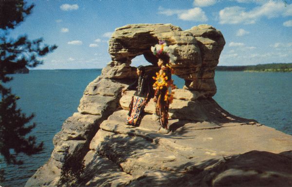 Text on reverse reads: "At Demon's Anvil, Wisconsin Dells, Wis." A Native American woman and man pose in front of Demon's Anvil wearing indigenous clothing. The Wisconsin River is in the background. Demon's Anvil is also known as Devil's Anvil.