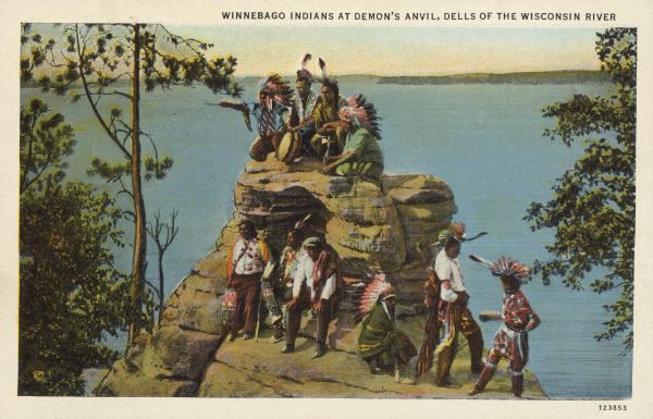 Text on front reads: "Winnebago Indians at Demon's Anvil, Dells of the Wisconsin River." On reverse: "Winnebago Indians at Demon's Anvil. Each summer some of the Winnebago Indians go into camp on the high land above Stand Rock, and near by they hold Ceremonial dances. This is a group of these Indians in gala attire." A group of Ho Chunk Native Americans pose on a rock formation, with the Wisconsin River in the background. Demon's Anvil is also known as Devil's Anvil.