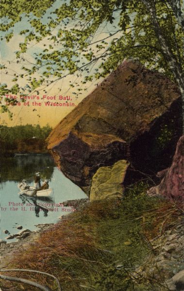 Text on front reads: "The Devil's Foot Ball, Dells of the Wisconsin." View from shoreline towards three people in a rowboat, floating near the Devil's Football, a rock formation on the shore of the Wisconsin River. The opposite shoreline is in the background.