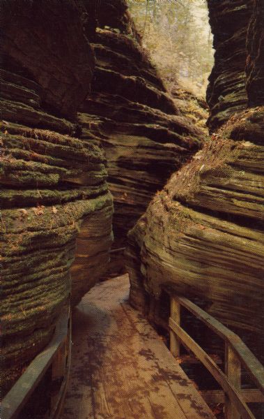 Text on reverse reads: "Fat Man's Misery. Through much of Coldwater Canyon the rock walls are very high and the passage between them is very narrow. The narrowest point is called Fat man's Misery for obvious reasons. Wisconsin Dells, Wis." A boardwalk in a narrow passage between two rock formations.