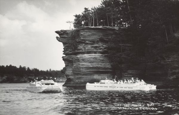 Text on front reads: "At the Hawk's Bill, Wisconsin Dells, Wis." Tour boats filled with sightseers on the Wisconsin River. Trees are clinging on the side of the formations, and are also growing on top. Text on the right hand boat reads: "See the World Famous DELLS by Land and Water."