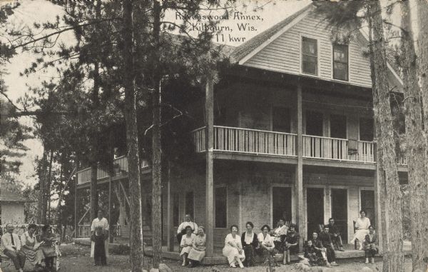 Text on front reads: "Ravenswood Annex, Kilbourn, Wis." A group of visitors poses on the porch and yard at the Annex at Ravenswood, a three-story building with a porch and a balcony above. Pines trees are on the grounds, and there is a smaller building in the background on the left.<p>Kilbourn City was founded in 1857 by Byron Kilbourn, the name was changed in 1931 to Wisconsin Dells.</p>