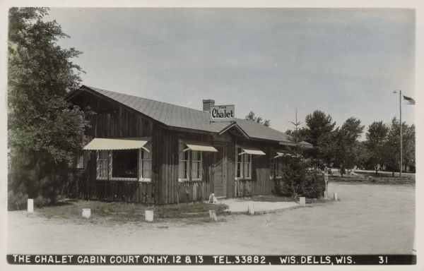 Text on front reads: "The Chalet Cabin Court on Hy. 12 & 13, Tel.33882, Wis. Dells, Wis." The log cabin office at the Chalet Cabin Court. A dog is sitting in front of the door.