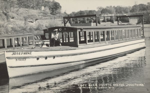 Text on front reads: "'Capt. Herb Droste' and the 'Josephine,' Wisconsin Dells, Wis." A tour boat and the captain, moored at the Dells Boat Company dock with other boats. The boat registration number is "37A333." Rock formations, trees and the dam are in the background.