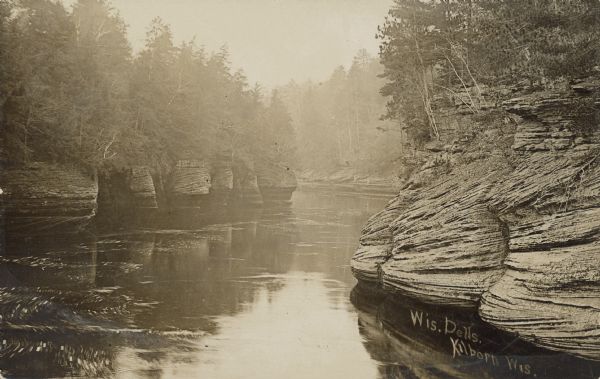 Text on front reads: "Wis. Dells, Kilbourn, Wis." Rock formations and trees line the shores of the Wisconsin River through the Dells.<p>Kilbourn City was founded in 1857 by Byron Kilbourn, the name was changed in 1931 to Wisconsin Dells.</p>