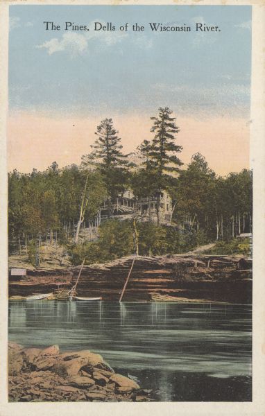 Text on front reads: "The Pines, Dells of the Wisconsin River." View from rocky shoreline towards the lodge at The Pines Resort, perched above the shore behind trees. Below the formations, at the river's edge, is a dock with rowboats.