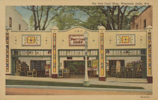 Text on front reads: "The Post Card Shop, Wisconsin Dells, Wis." On the reverse : "The Post Card Shop is situated across from the boat docks. It is a convenient place to shop, write postcards or rest while watching the passing parade." View of the storefront, which is filled with postcards and other souvenirs. The building has three large openings and transom windows. There are Native American inspired motifs decorating the front. On each side are commercial buildings. Trees are in the background.