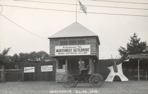 Text on front reads: "Wisconsin Dells, Wis." A model historical village with a guardhouse flying the American flag in the center, flanked by fences and more buildings. A vintage automobile is parked in front, with a tipi on the right. In the background are trees. The three signs read: "Entrance to- Northwest Settlement, Model Historical Village", "See Fawn Deer Exhibit, Bottle Fed Every 2 Hrs." and "See the Old Saloon, Jail, Fire Station, Blacksmith Shop, Livery, Store, Church."