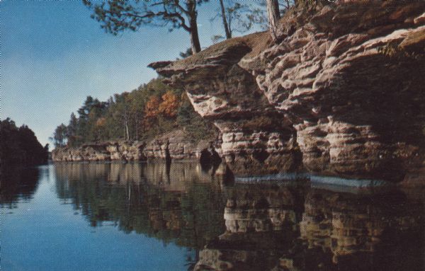 Text on reverse reads: "Swallows' Nests, Wisconsin Dells. Every summer hundreds of swallows make their homes in holes in the soft sandstone cliff." Rock formations on the shore of the Wisconsin River with trees growing above.