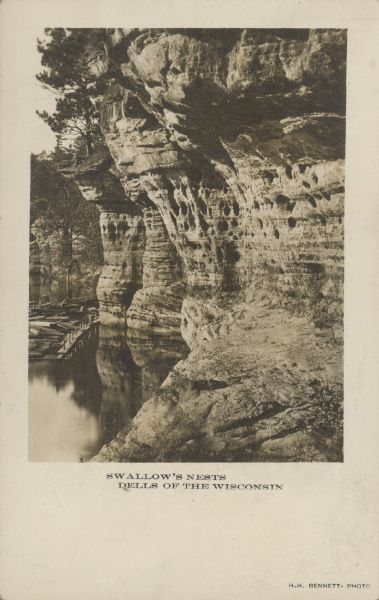Text on front reads: "Swallow's Nests, Dells of the Wisconsin." Rock formations on the shore of the Wisconsin River with trees growing above. Every summer hundreds of swallows make their homes in holes in the soft sandstone cliff. A lumber raft is at the base of the cliff on the left.
