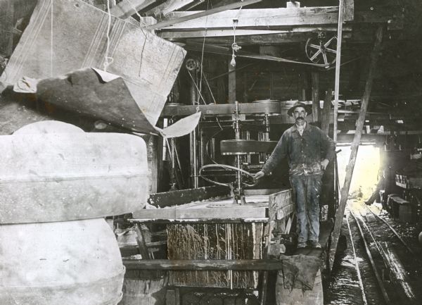 A workman poses beside a belt-driven polisher inside a shed at the Montello granite quarry. Two other men relax in the background near an open door where narrow gauge tracks enter the shed.