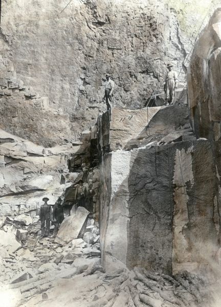 Two men stand on a ledge in the granite quarry in Montello. Three other men stand below. The side of the quarry rises up above the group in the background. Several bore holes are apparent on the cut faces of the stone.