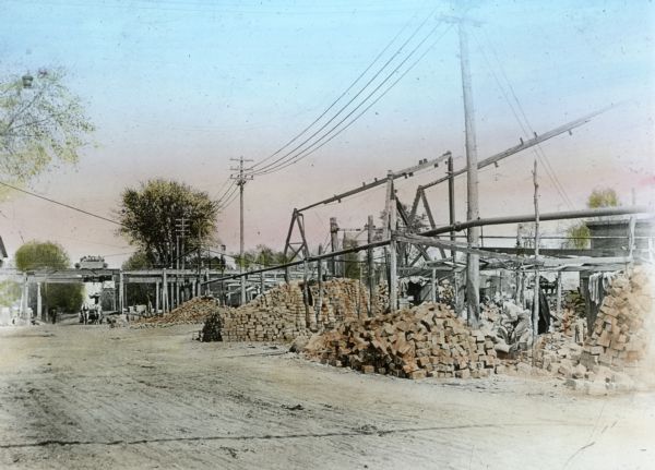 View of the Montello granite quarry grounds shows piles of granite paving blocks. In the background, a team of horses pulls a wagon under an elevated narrow gauge railway as a rail car crosses above. There is a worker among the granite paving blocks in the foreground on the right.
