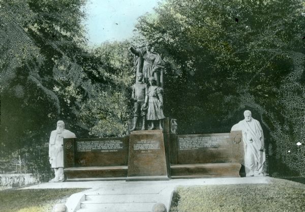 Hand-colored lantern slide of the Illinois Memorial at Andersonville National Cemetery. A bronze sculpture depicting Columbia, Youth and a Maiden is mounted on the central pedestal of the wide base, which is made of Montello granite. On each side is a sculpture depicting a weary Civil War veteran. The sculptures are also of Montello granite.  Although the slide depicts the side sculptures as white, they are actually light pink. The cost of the granite work was $15,000.