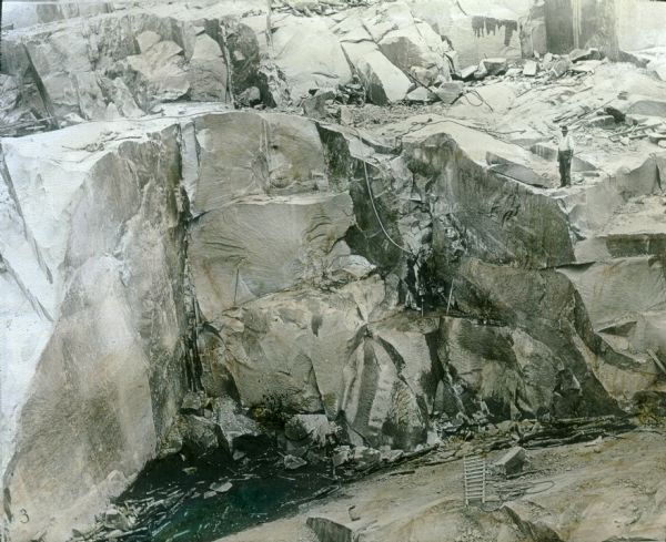 Elevated view looking southeast at a cut face of the granite quarry. A worker stands on a ledge on the right. There are ropes and ladders in the quarry.