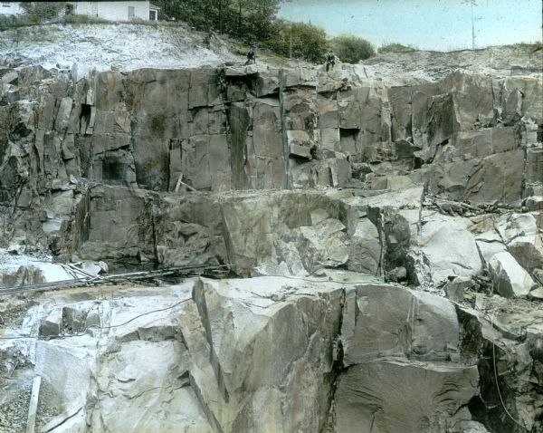 View over quarry towards two men in suits sitting at the top edge of the cut face of the granite quarry. In the background is a house at the top of the hill near the edge of the quarry.