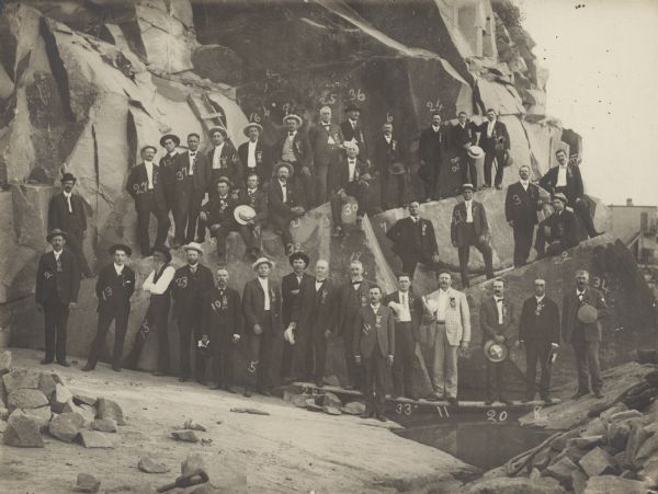 Officers, sales representatives and clients of the Montello Granite Company posing in the quarry. Most of the men are wearing ribbons pinned to their jackets. The printed key to the identification of the men is mounted on the reverse of the photograph.