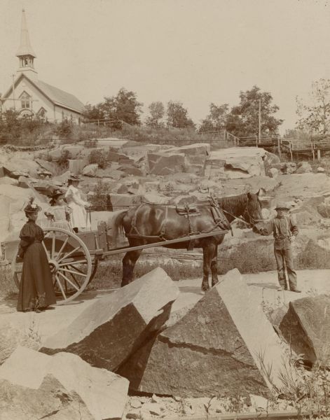 A boy is standing and holding the bridle of a horse harnessed to an open wagon in the granite quarry. Ethel Richter, standing in the wagon with her sister Blanche, is holding the reins. The girls' mother, Mary Richter, is standing beside the wagon. A wooden walkway leads to the Catholic church at the top of the hill. Mary's husband, Charles Richter, was an owner of the Montello Granite Co.
