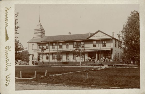 A cabinet card photograph of the Montello House Hotel. The two-story brick building has a gable fronted wing on the right and a shingled wooden tower on the left which held a water tank. The two-story porch extends along the entire front. There is an electric light hanging from a pole in front of the hotel. People are posing on the lower porch on the right; the man on the left is wearing a chef's uniform. A sidewalk crosses the lawn. On the reverse is written: "The hotel where we boarded in 1897."  