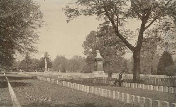 A man is sitting on a headstone near the Iowa monument at the Andersonville National Cemetery. He is holding an umbrella in his right hand, and his hat and coat in his left. The New Jersey monument is toward the left; other monuments are visible in the background. The dark pedestal of the Iowa monument, which supports a statue of a weeping woman, is made of red granite quarried in Montello, Wisconsin.  