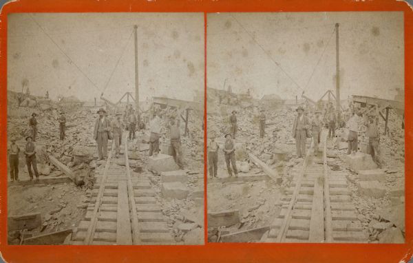 Stereograph of a bearded man with vest and suit posing on a narrow gauge railroad at the granite works in Montello. Workmen are posing around him. Most of the men are facing the camera, but two men on the right are holding mauls overhead, appearing to work on large blocks of granite. There are buildings and two derricks in the background.
