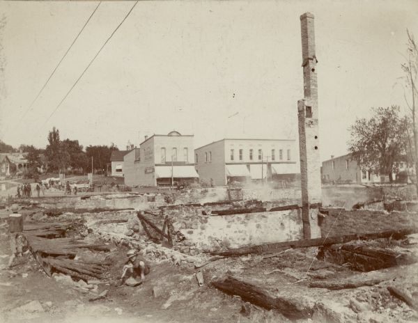 Elevated view of two barefoot children who are near a charred wooden sidewalk, lower left, that marks the course of Nebraska (now West Montello) Street toward its intersection with Main Street, where a small crowd has gathered. Buildings which once faced Nebraska Street have been destroyed by fire. To the right, smoke is rising from the basements of buildings that fronted Main Street and were destroyed by fire. An unsupported chimney is standing in the foreground.  