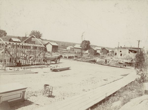 Elevated view of a horse-drawn wagon loaded with lumber standing in front of the frame of building under construction facing Main Street. There is a worker on the wagon and two others at the building site. In the foreground is the wooden sidewalk along Barstow (now East Montello) Street; a store counter and chair are sitting on the bare ground facing the sidewalk. There are open basements along Main Street, and visible in the background, right, is the side wall of a commercial building damaged by fire. 